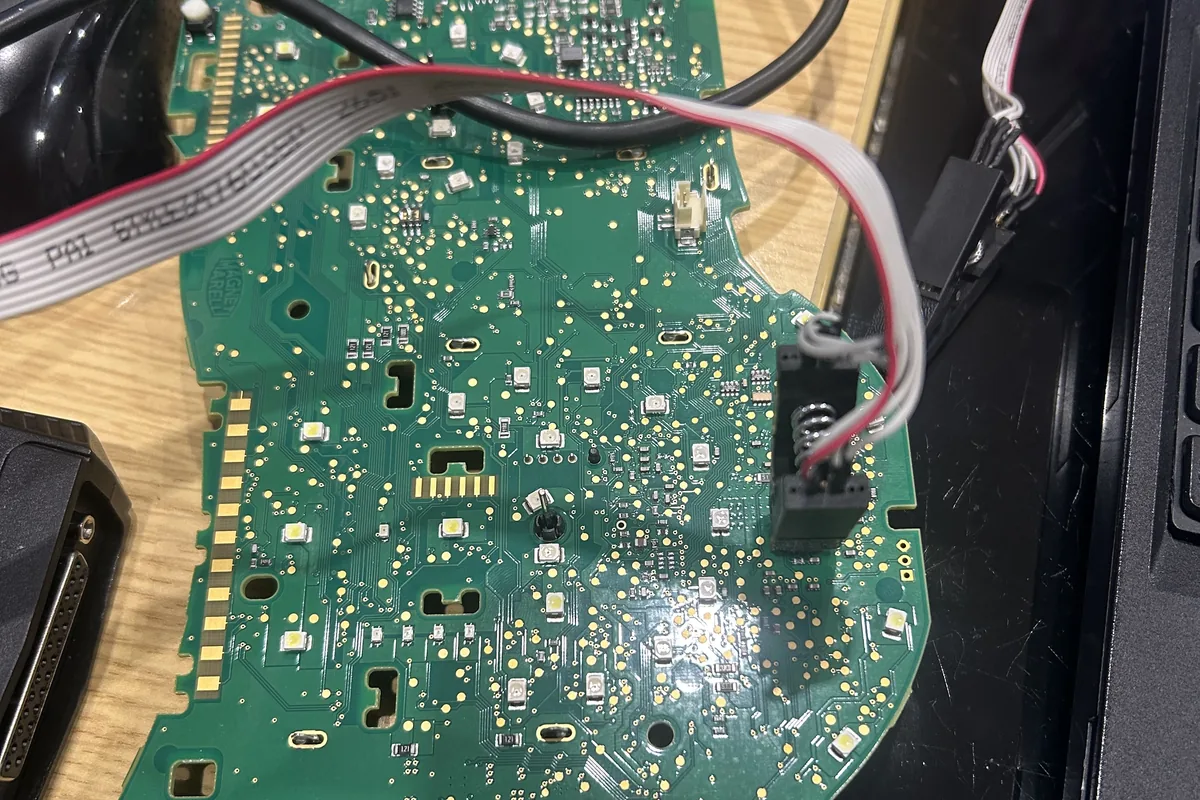 A Porsche instrument cluster board on the bench with a clip on the memory chip and a ribbon lead to the programmer